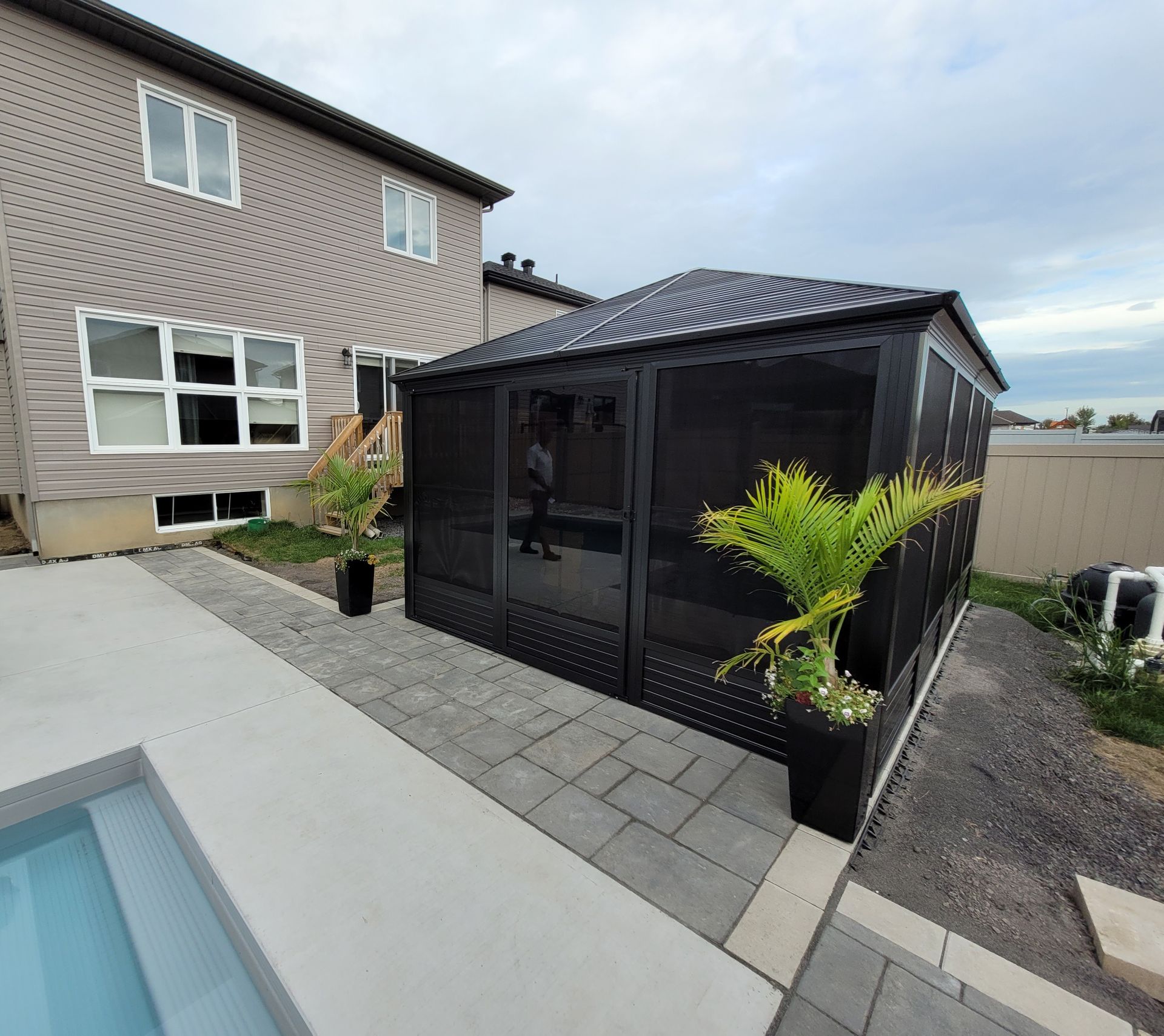 Black screened gazebo next to a house and a pool. Features plants and a paved walkway.