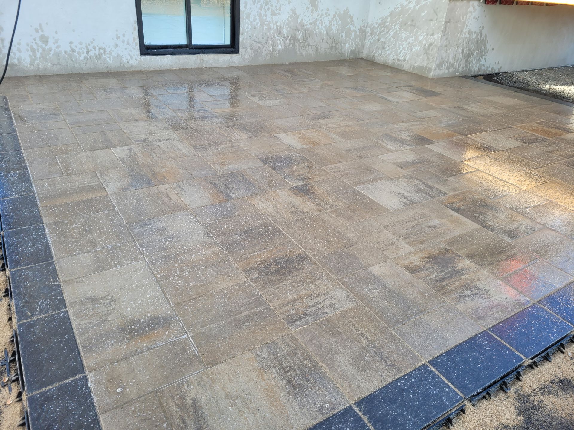 Brick patio with dark border, wet from rain, next to a light stucco wall and window.