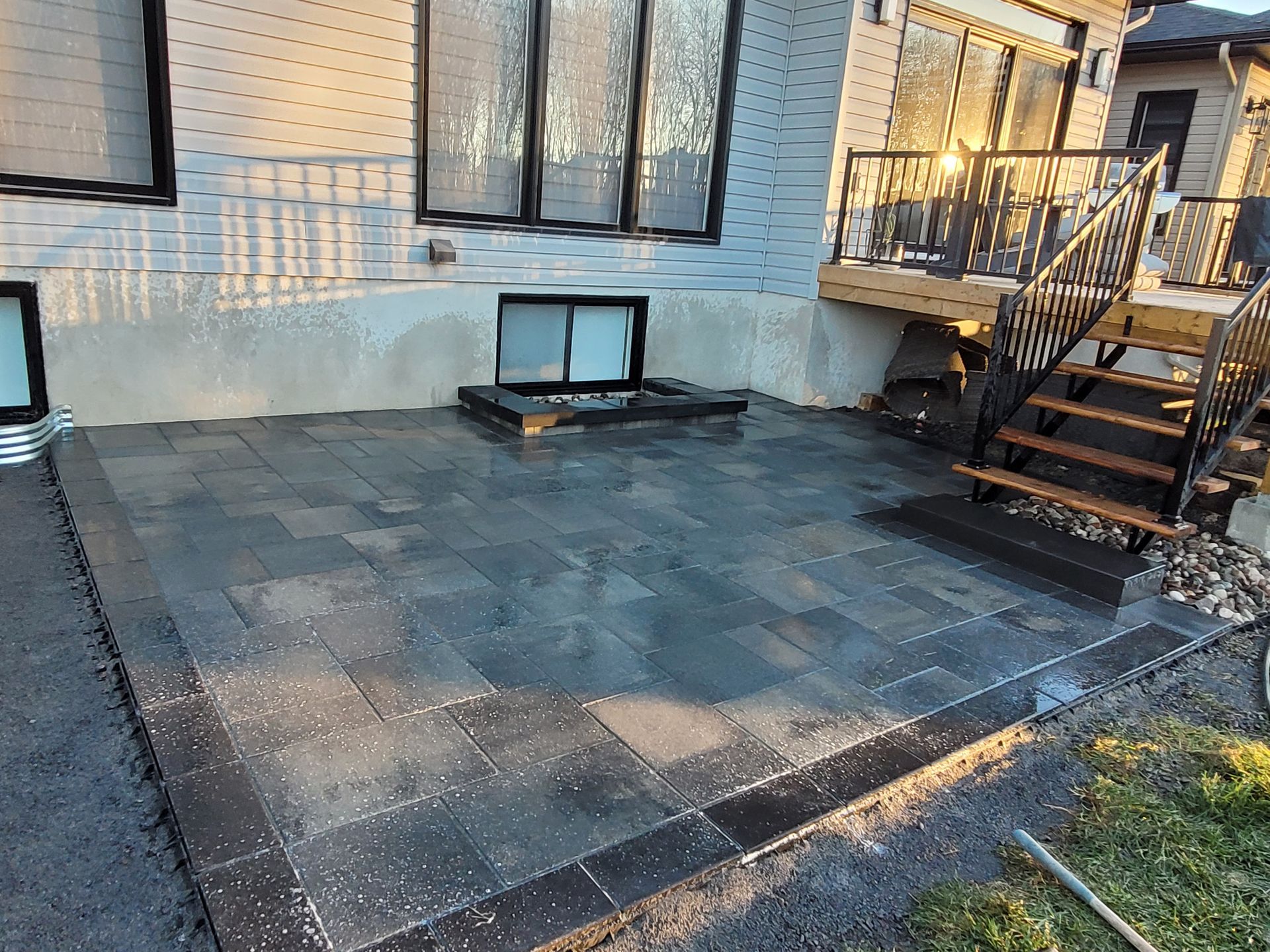 Patio with dark gray pavers bordered by a darker edge. A house with windows and a deck are visible.