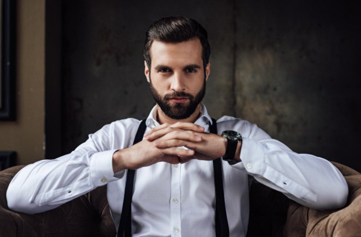 Un hombre con barba y tirantes está sentado en un sofá con las manos cruzadas.