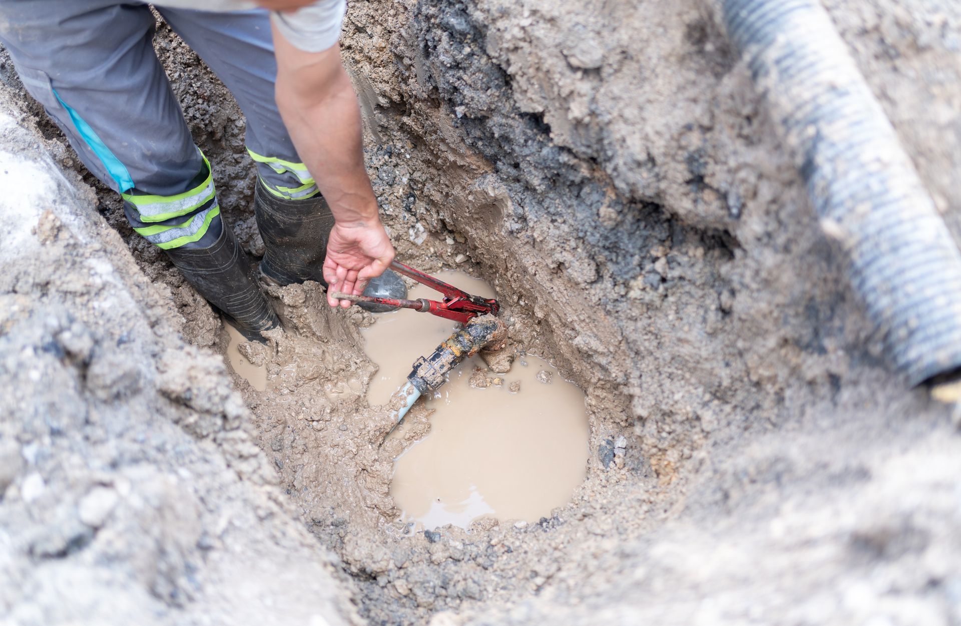 Plumber repairing a broken underground water pipe with adjustable wrenches and tools at roadside.