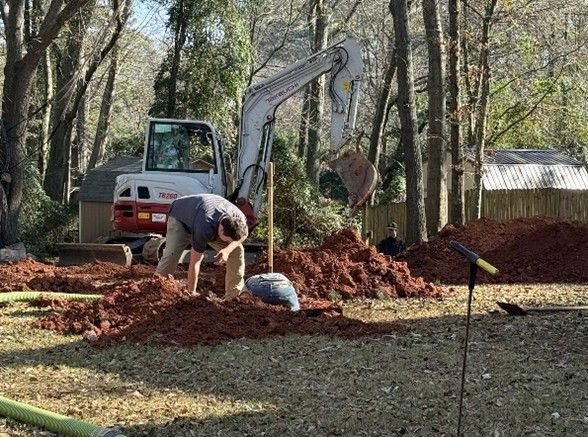 Man working on the ground with an excavator in a yard, digging. Red soil, trees, and cloudy sky. Man working on the ground with an excavator in a yard, digging. Red soil, trees, and cloudy sky.
