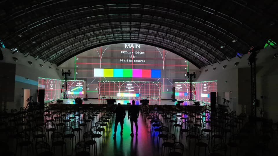 Ampio spazio al coperto con palco e schermi che mostrano barre colorate. Due figure sono in piedi davanti al palco.