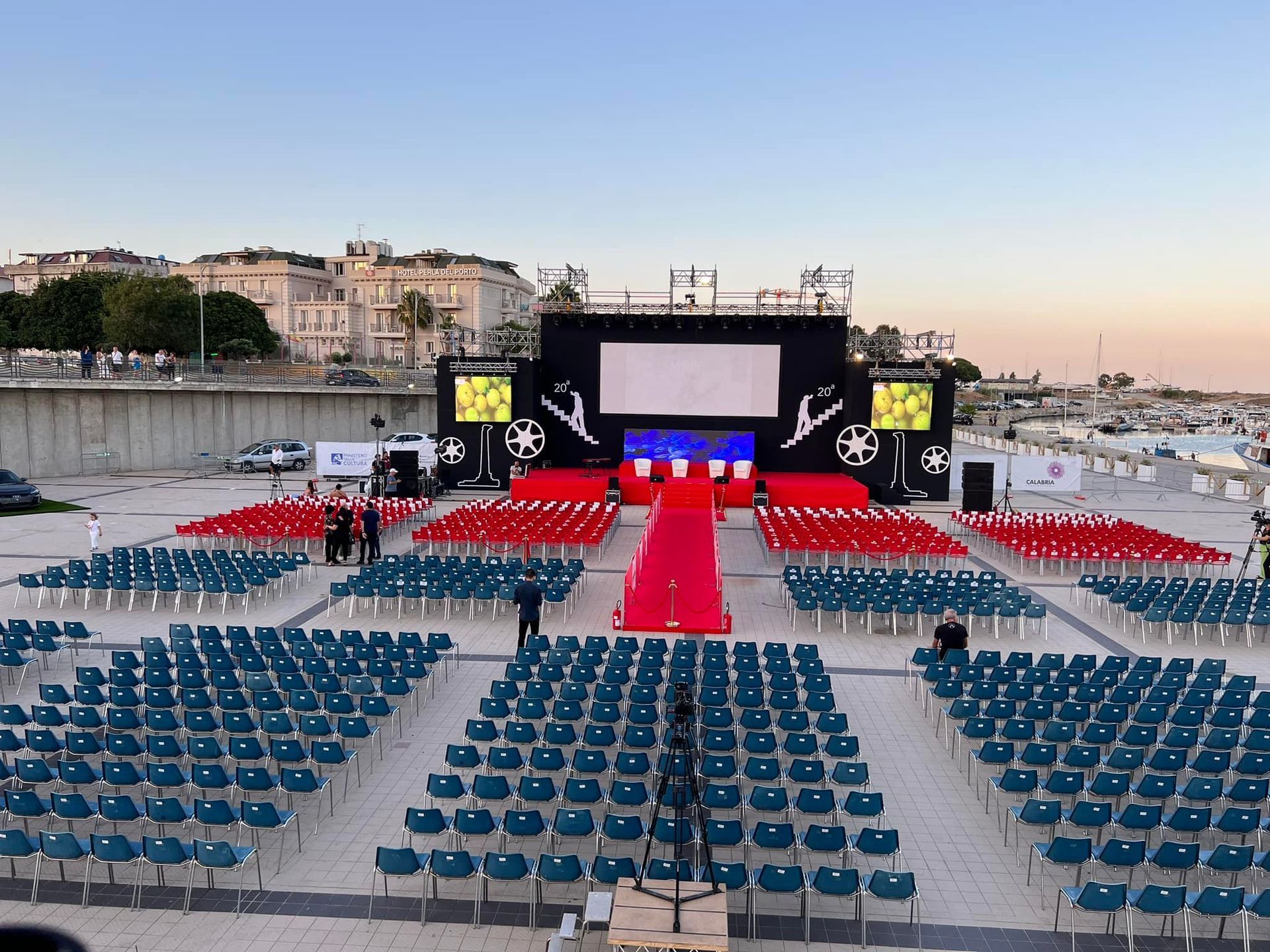 Allestimento di schermi cinematografici all'aperto; file di sedie blu e rosse, palco e fondale del porto.
