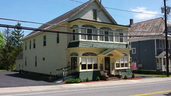 Two-story yellow building with green trim, arched windows, and a black paved driveway on a sunny day.