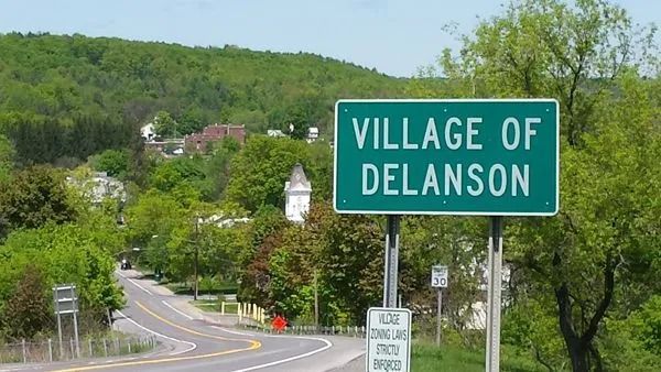 Village of Delanson sign along a road with a church in the background, green and sunny day.