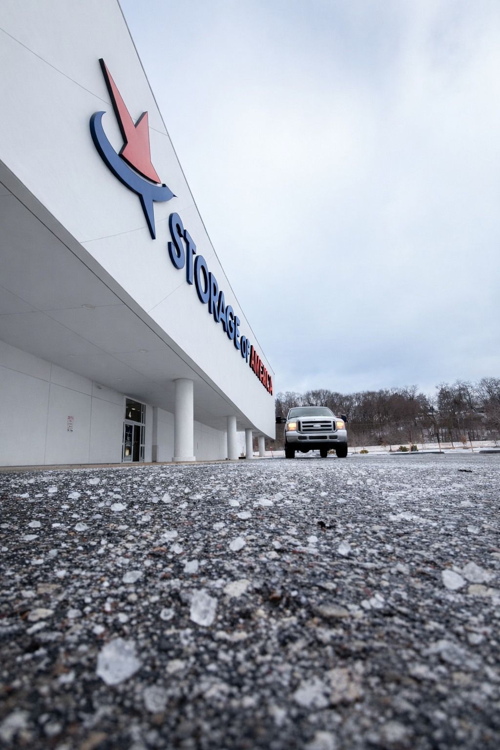 Low-angle view of a Storage Mart building with a white facade and blue and red logo, with a parked truck in the distance.
