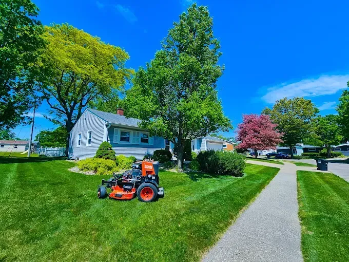 A bright orange commercial lawn mower parked on a freshly cut, manicured lawn in front of a house on a sunny day.