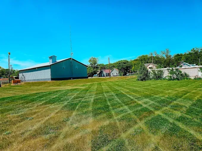 A large, sunny lawn mowed with a precise crosshatch pattern next to a long metal building and houses under a blue sky.