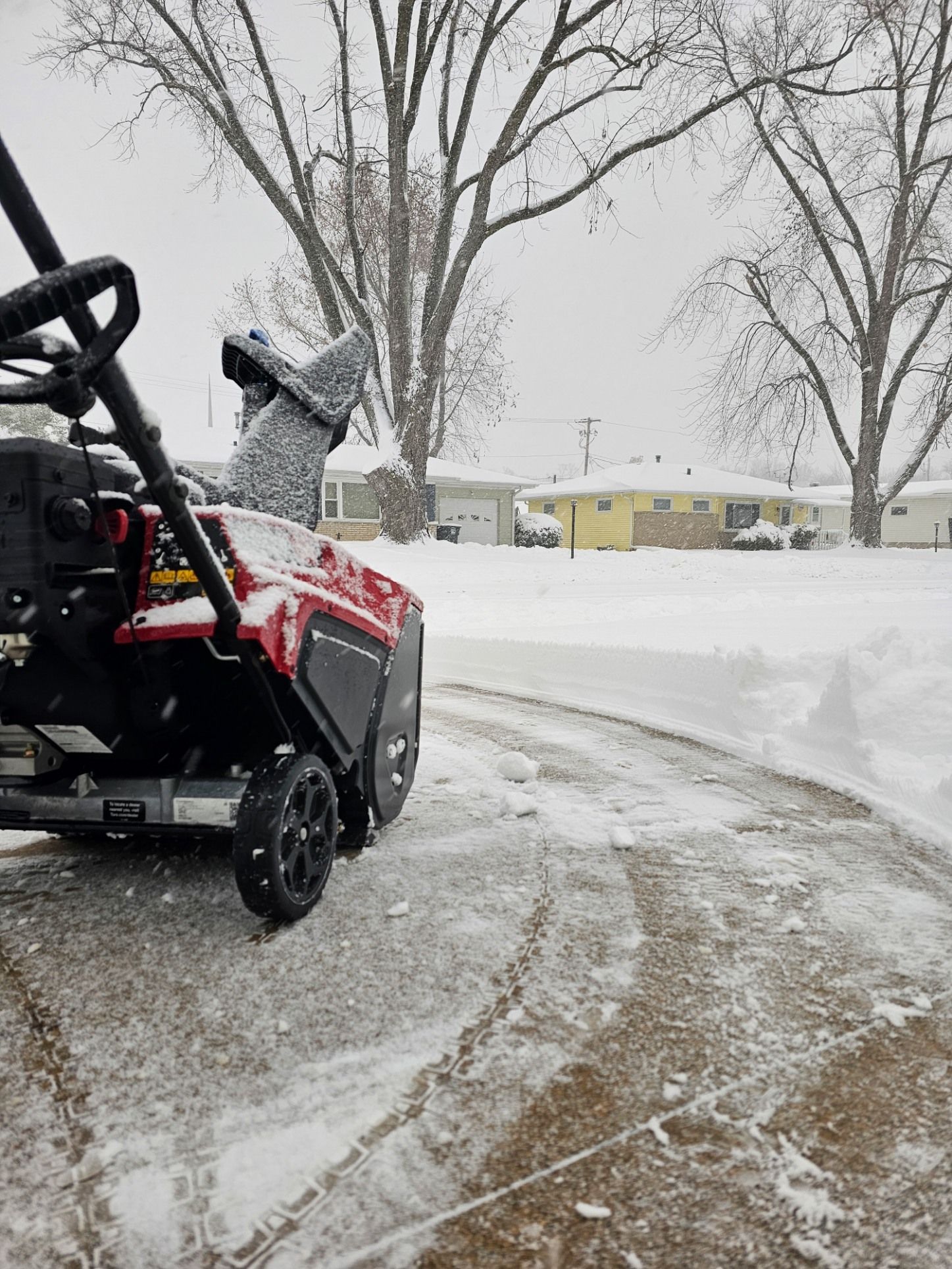 A red snowblower sits on a partially cleared suburban driveway during a snowy day, with houses visible in the background.
