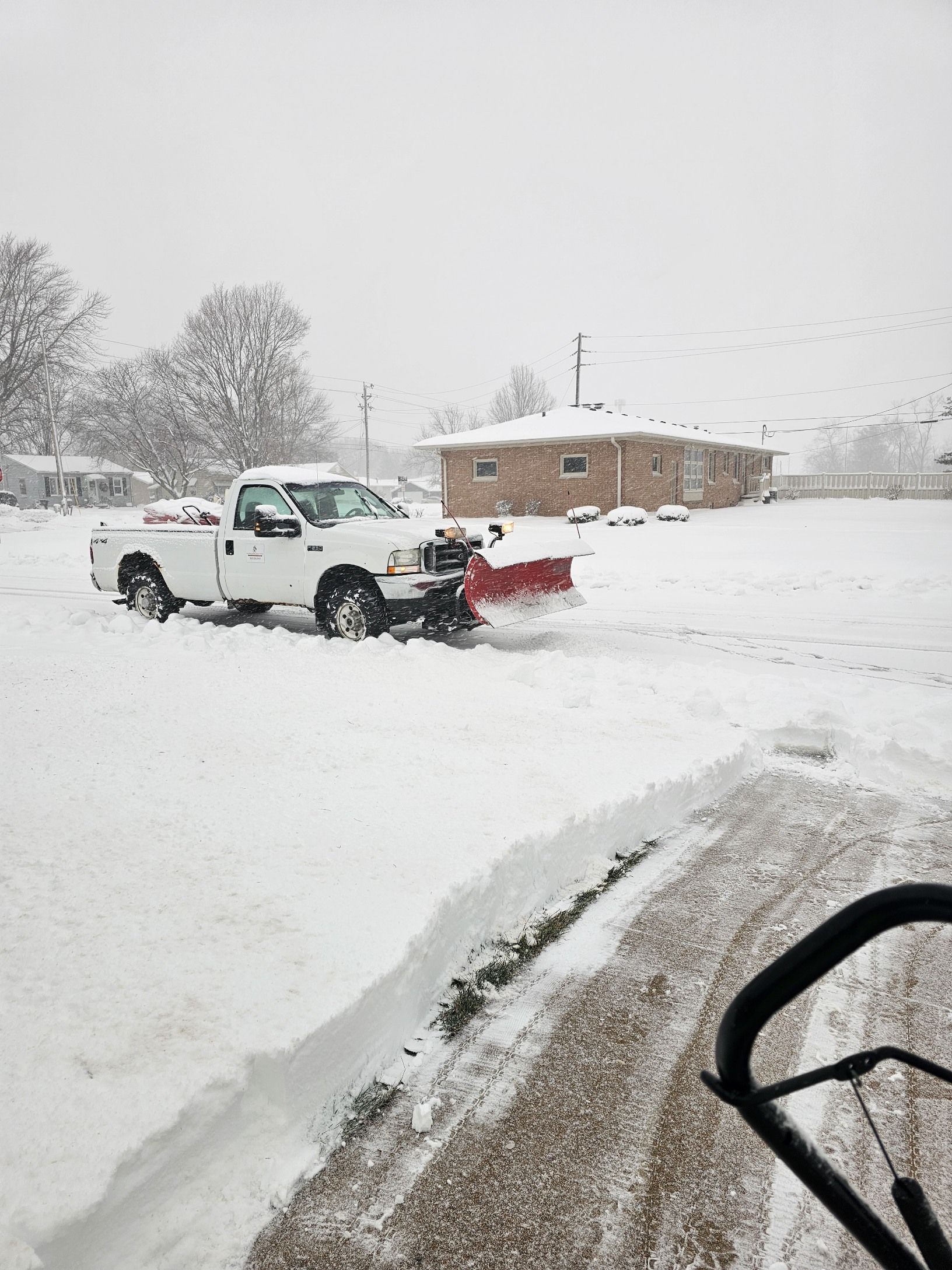 A white plow truck clears snow in a parking lot on a snowy, overcast day, viewed from behind a snowblower.