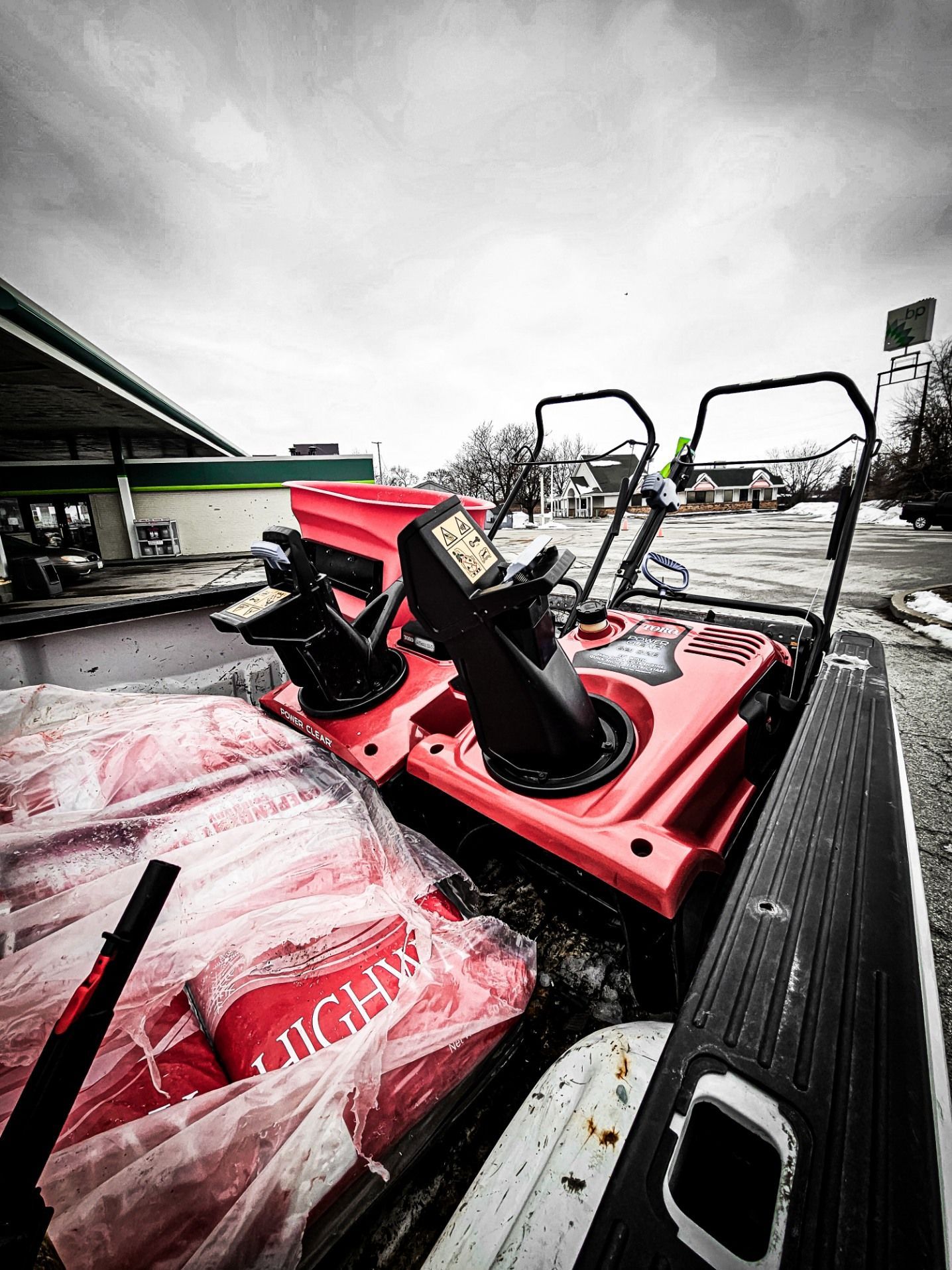 A red snow blower and bags of rock salt in the bed of a pickup truck in a parking lot on an overcast day.