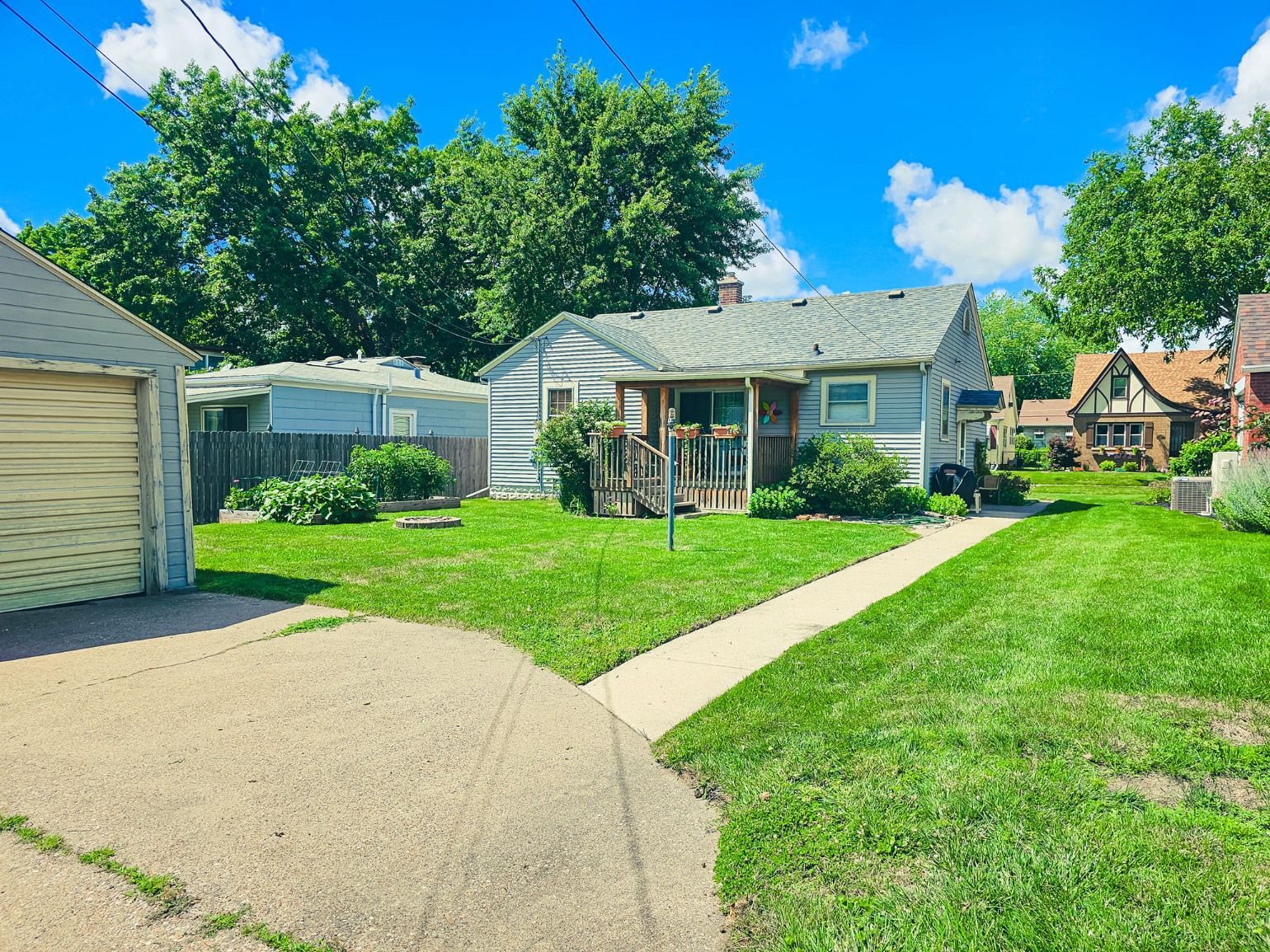 A grey single-story house with a wooden porch and a detached garage sits on a grassy lot under a bright blue sky.
