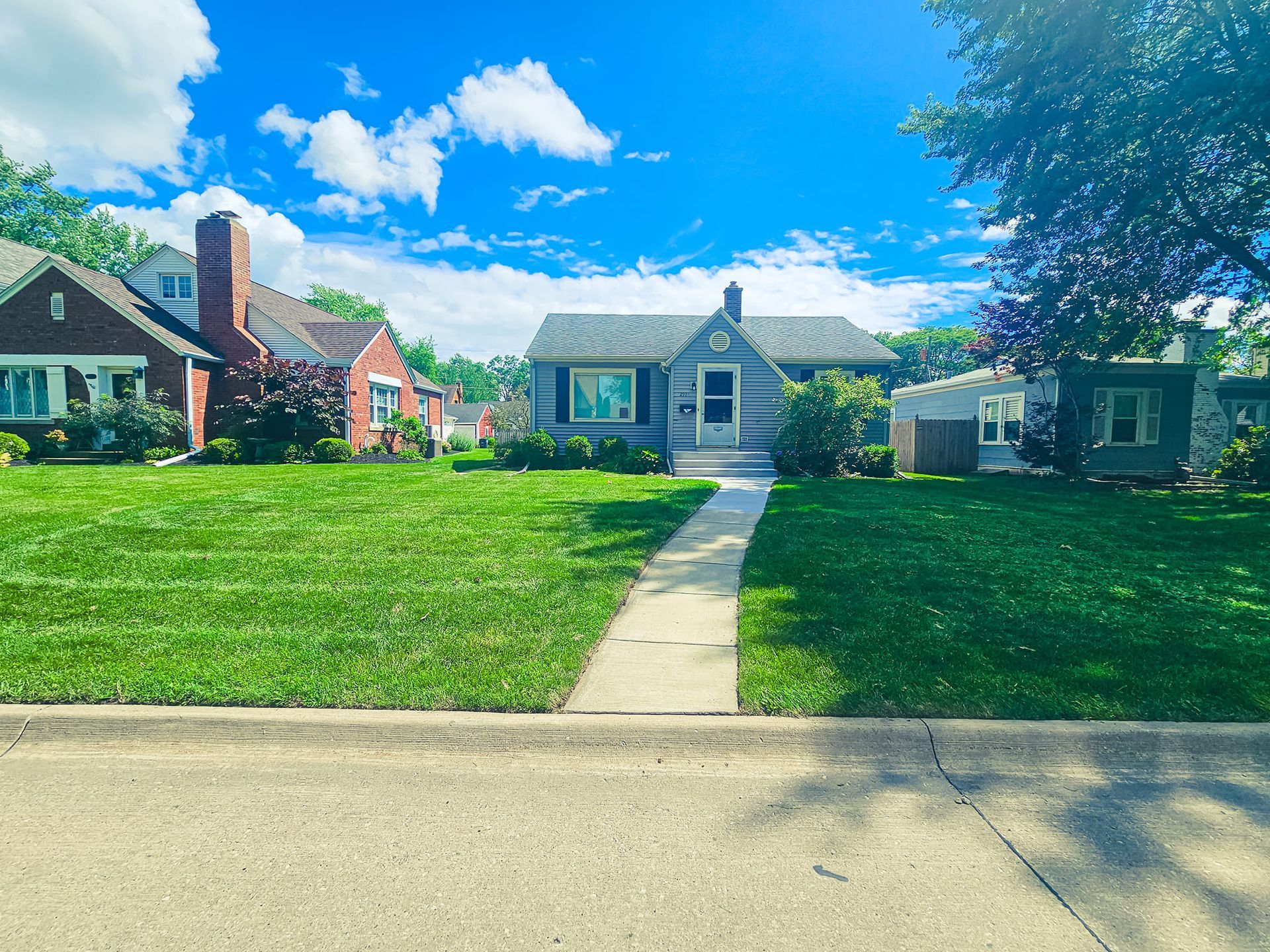 A residential neighborhood with a blue house, a brick house, and a grey house, each with a front lawn and a sidewalk.