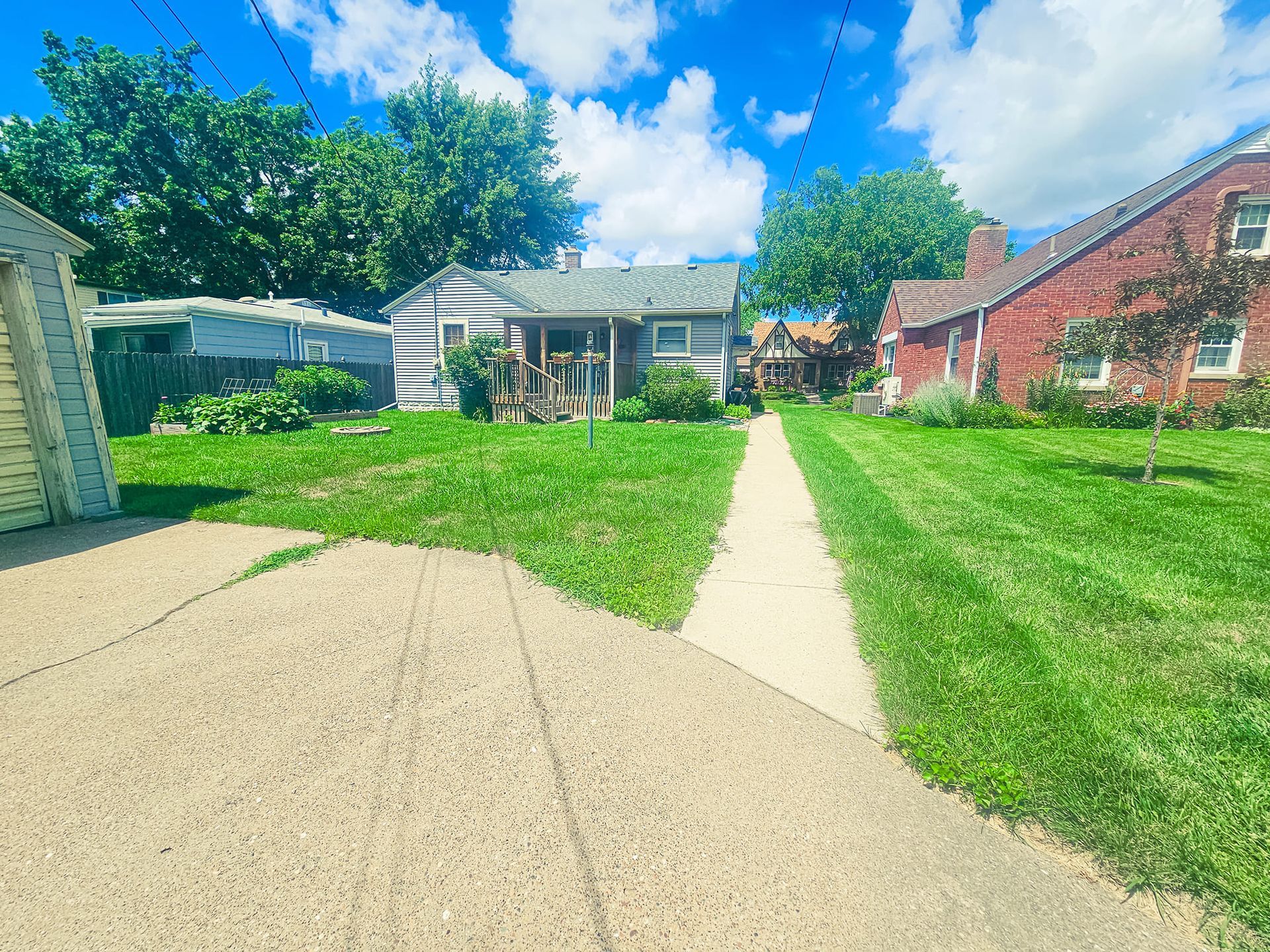 A residential backyard with a concrete path leading to a small house, flanked by green lawns and mature trees under a sky.