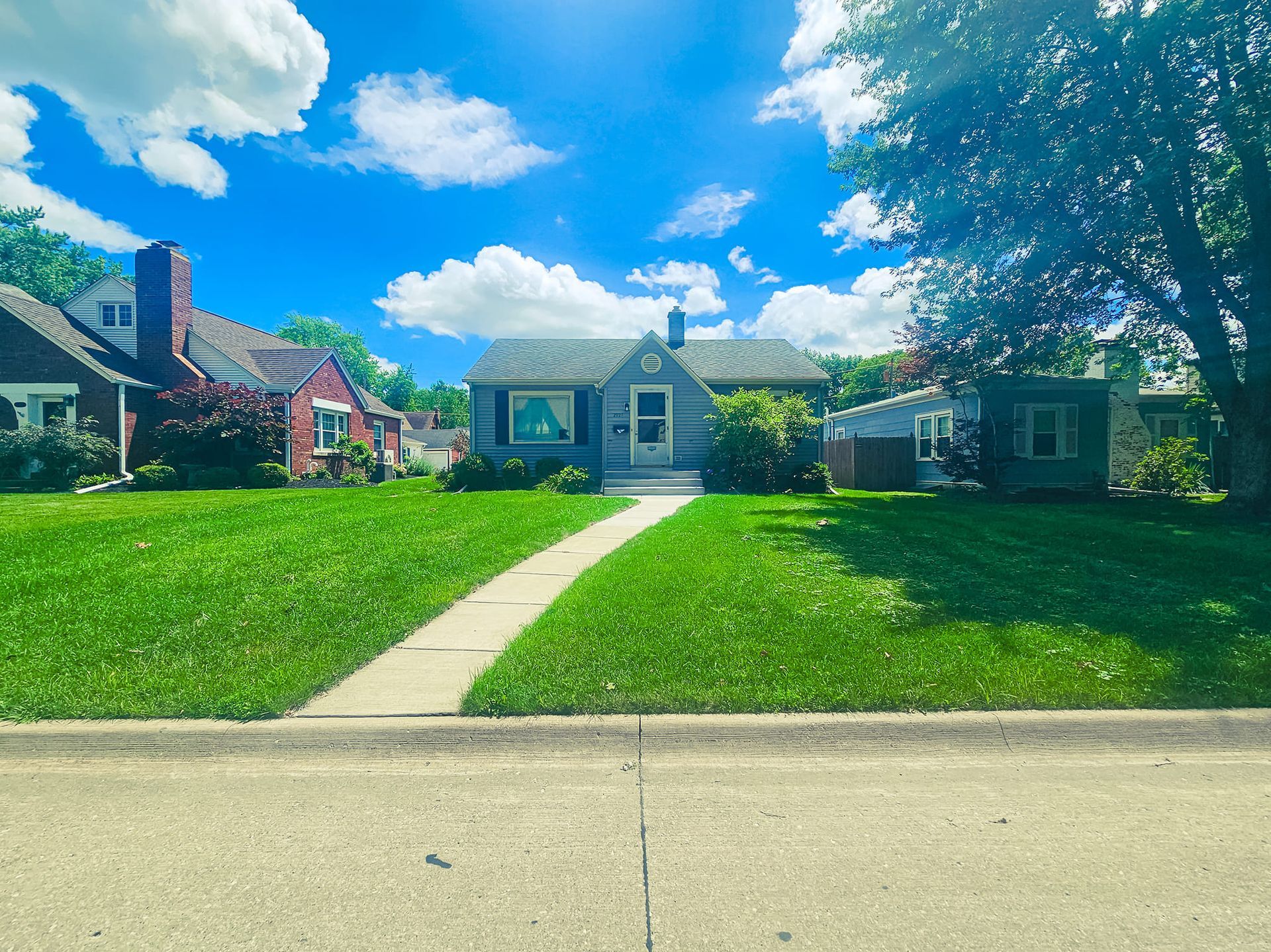 A street view of a small grey house with a white door and walkway, situated between two other homes under a blue sky.