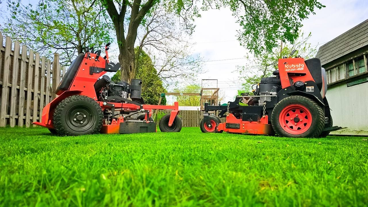 Two red stand-on lawn mowers sit facing each other on a lush green lawn in front of a wooden fence and house.