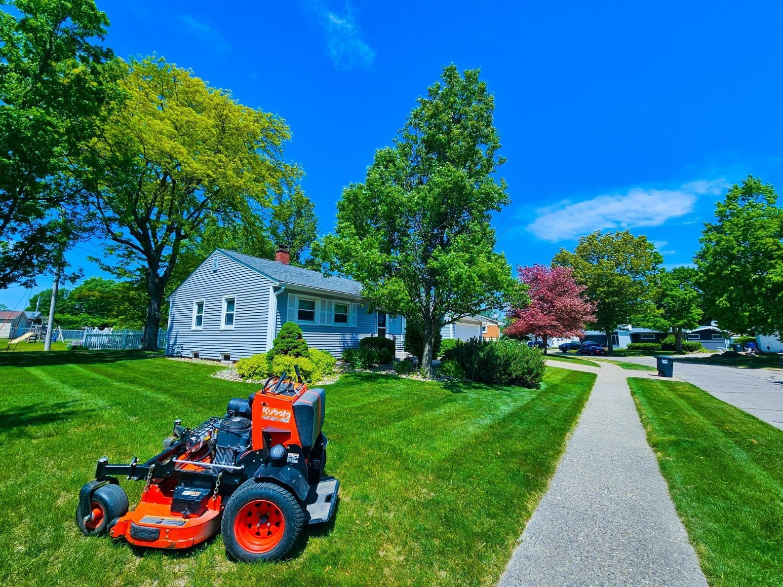 A bright orange lawn mower parked on a freshly cut, striped green lawn in front of a house on a sunny day.