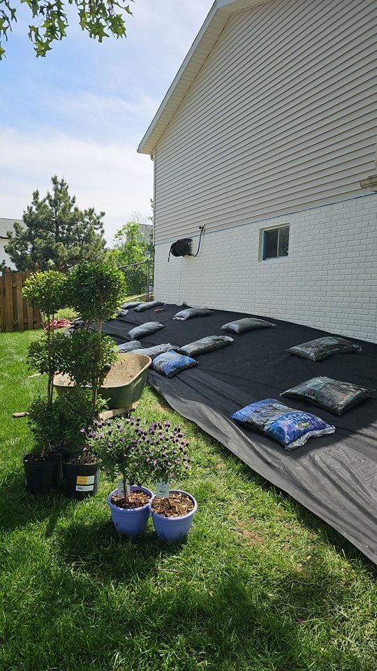 A row of potted shrubs sits next to a house foundation covered in black landscape fabric and bags of mulch.