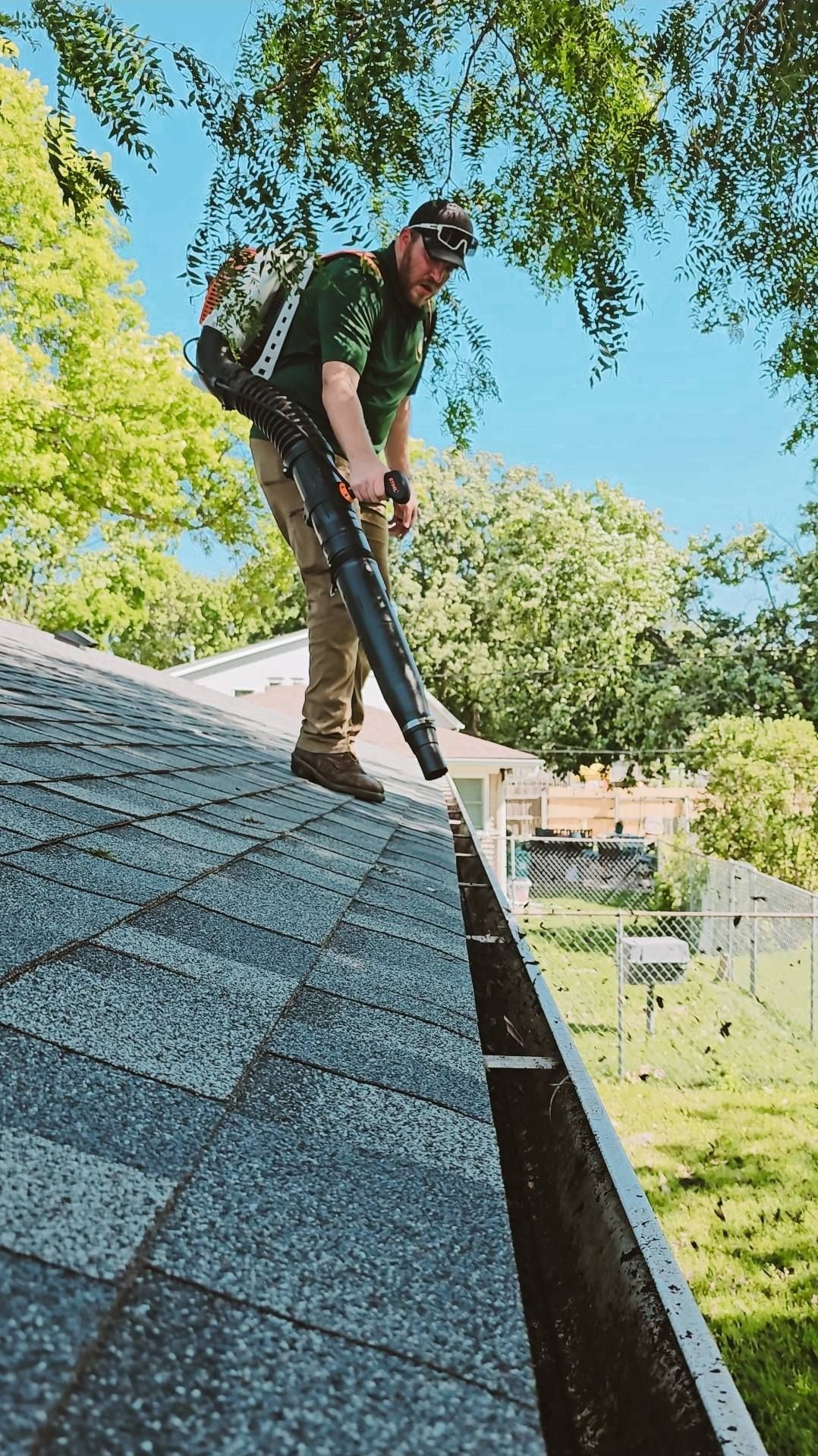 A person wearing a backpack blower stands on a slanted shingled roof, clearing debris from a rain gutter.