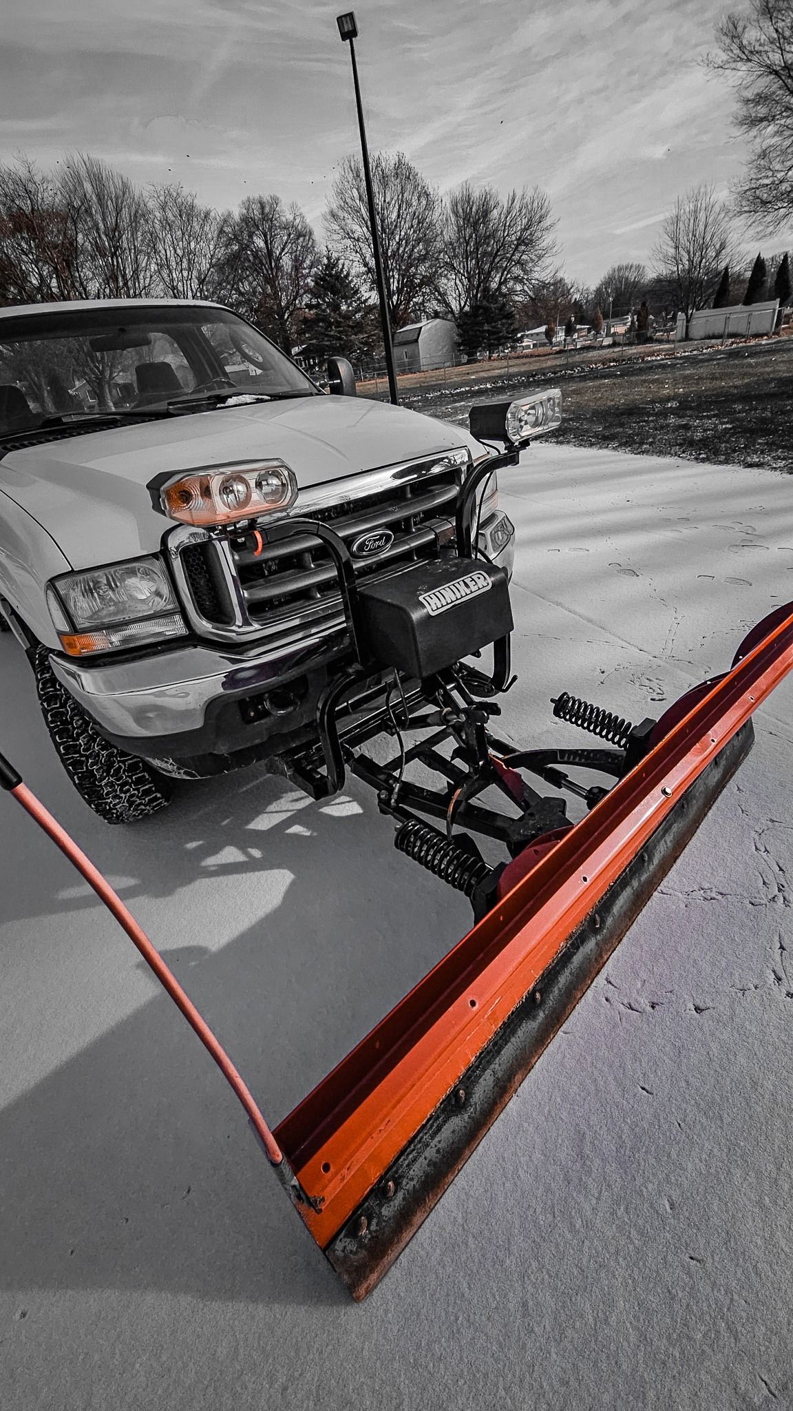 A light-colored pickup truck equipped with a large, orange industrial snow plow on its front bumper.
