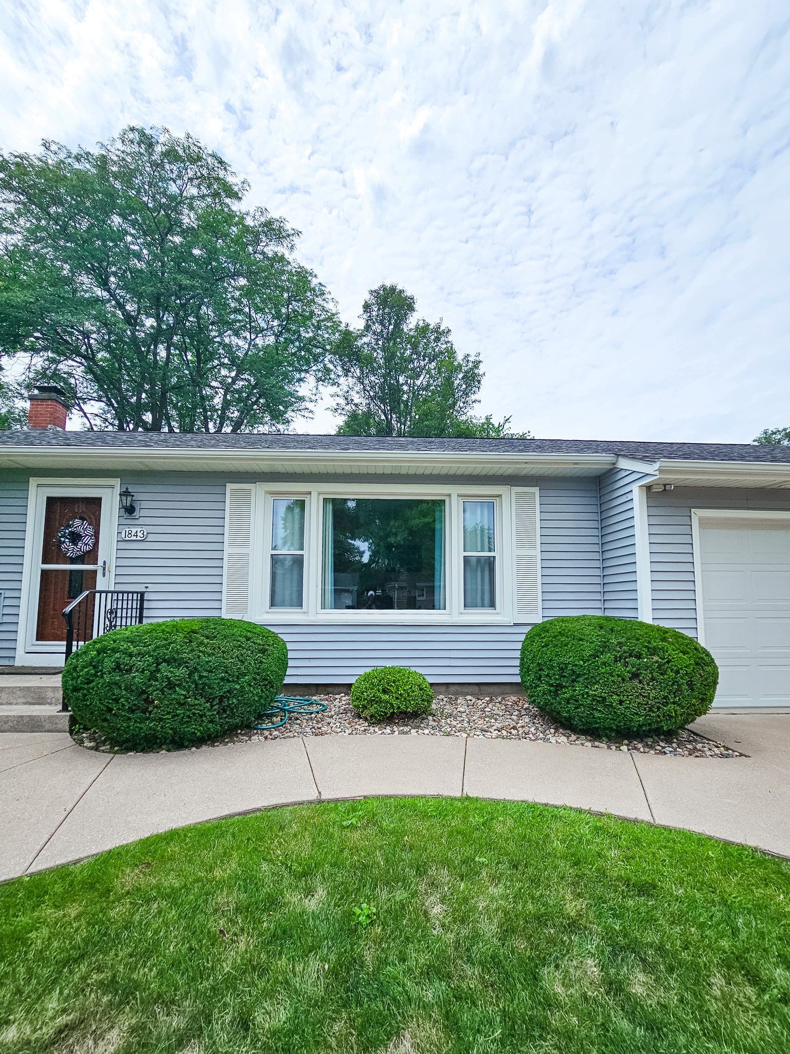 A light blue, single-story suburban house with white trim, a large front window, and a green lawn under a cloudy sky.