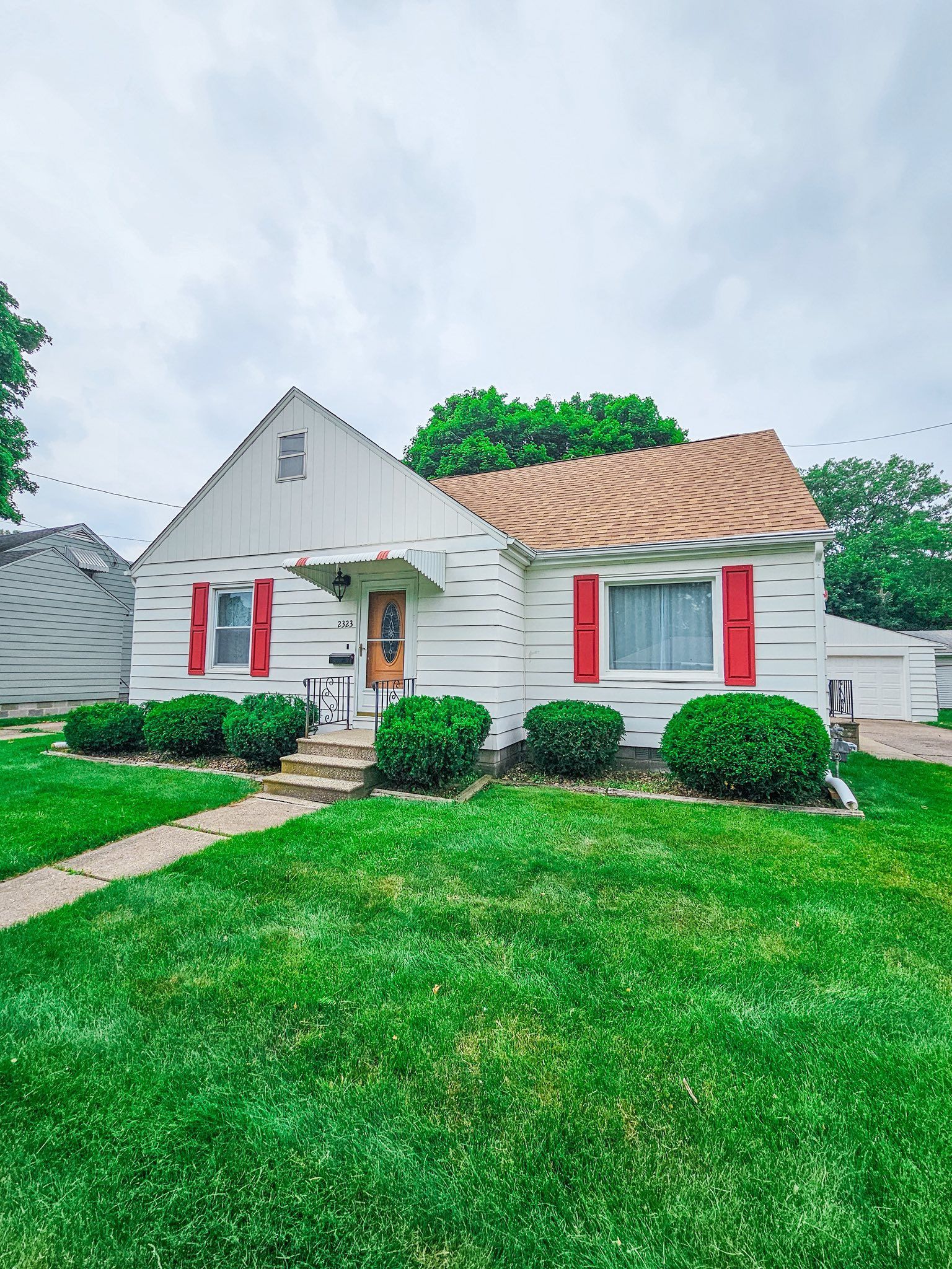 A white cottage with brown shingles and bright red shutters, featuring a lush green front lawn and a small walkway.