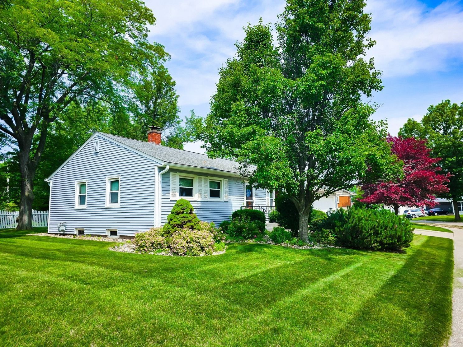 A light blue, single-story house with a grey shingled roof, set behind a neatly mowed lawn and mature trees.