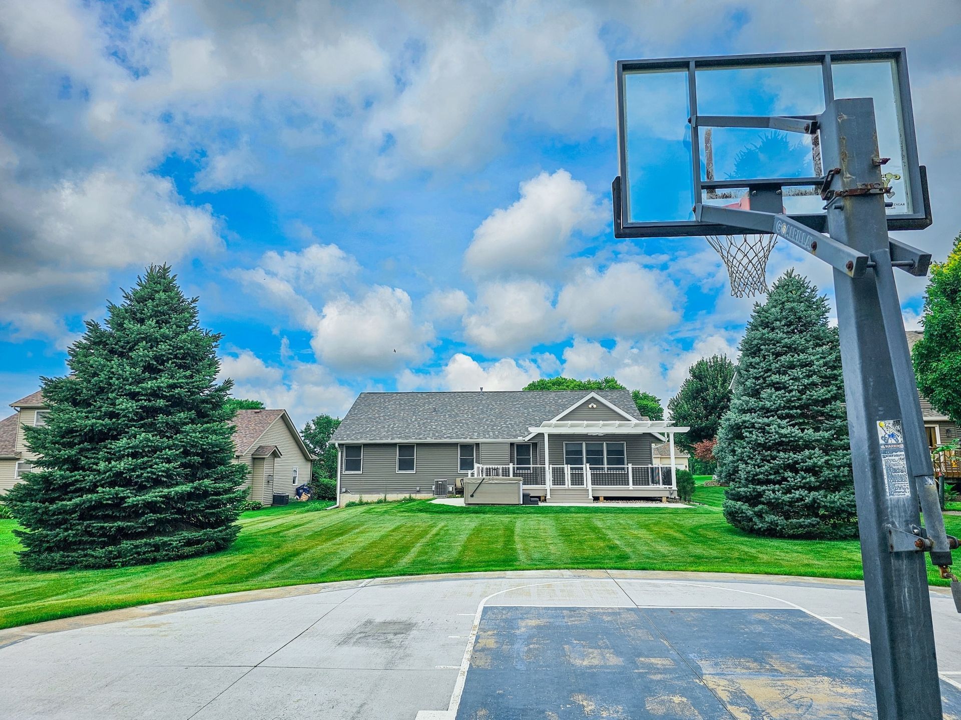 A basketball hoop stands in the foreground of a large suburban backyard with a gray house and green lawn under a blue sky.