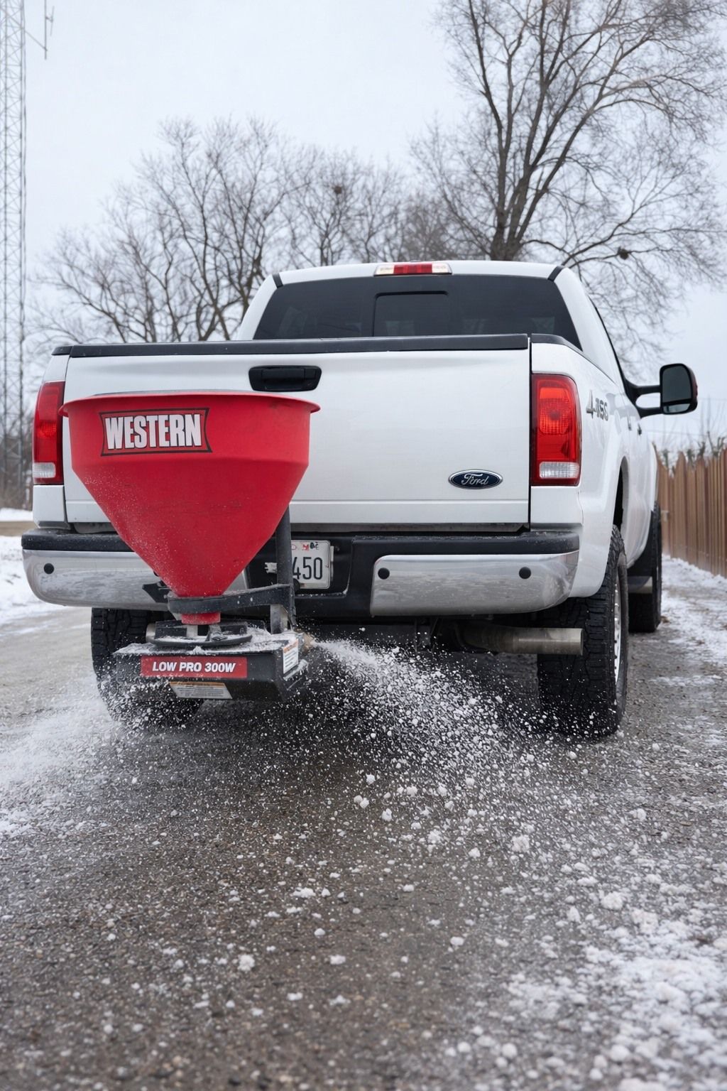 A white pickup truck with a red Western brand salt spreader mounted to the rear, scattering salt on a snowy road.