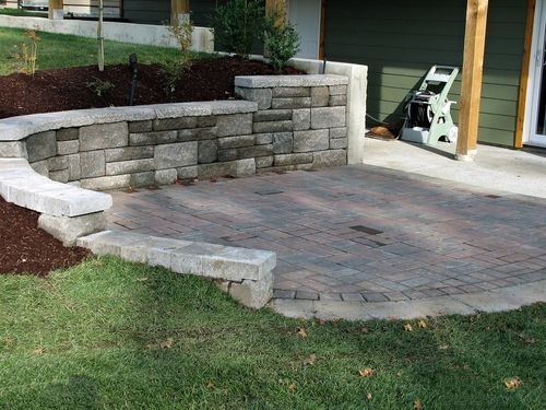 A curved stone retaining wall bordering a paved patio next to a house with green siding and a hose reel.