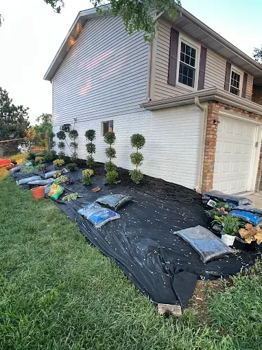 A suburban house side yard with topiary trees and bags of mulch placed on landscaping fabric for a garden project.