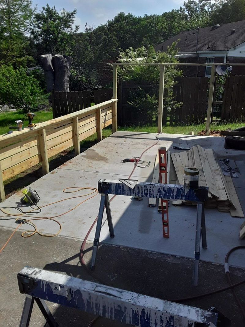 Construction site on concrete patio: fence with horizontal slats, pergola posts, sawhorses, lumber.
