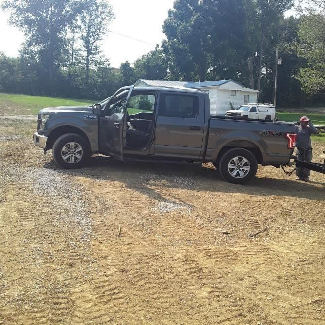 Gray pickup truck with door open, towing a trailer on a dirt lot. A person stands nearby.