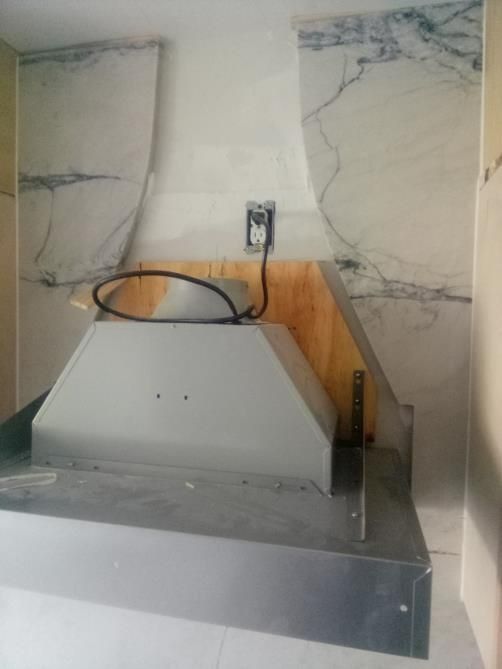 Stainless steel range hood under construction against marble tile backsplash. Electrical outlet visible.