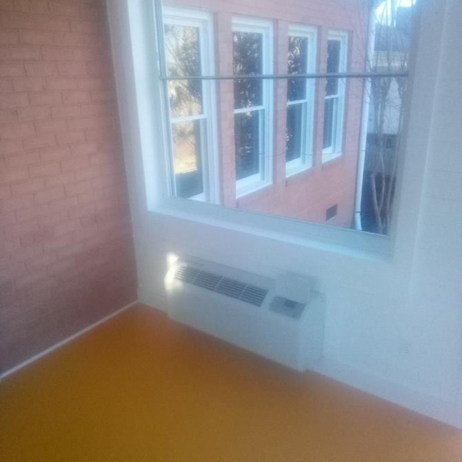 A room with a window showing a building, a wall with red bricks, a yellow floor, and a white air conditioning unit.