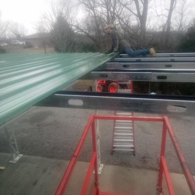 Person installing green metal roofing panels on a building. Red lift platform in foreground.