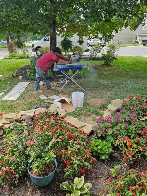 Person cutting stone with a saw outdoors, surrounded by plants and flowers. A white van is in the background.