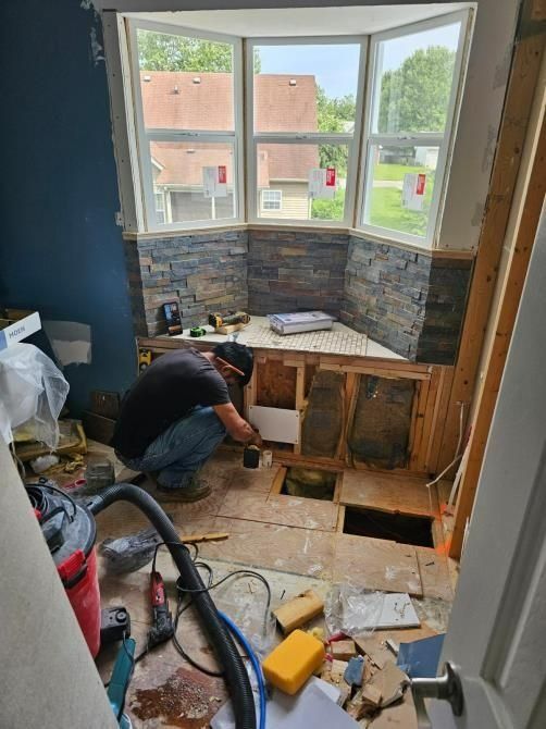 Person tiling a bathroom floor during renovation, near a bay window, with visible construction materials and tools.