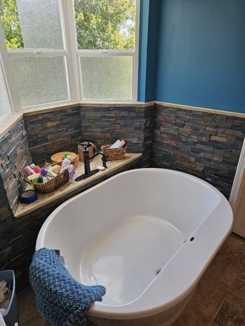 White oval bathtub in a bathroom nook with stone tile surround. Products on a built-in shelf, blue wall.