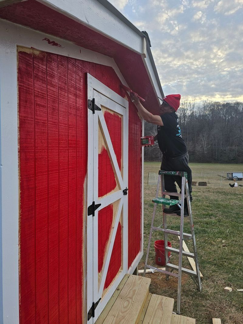 Person painting the red trim of a white-trimmed shed, standing on a ladder. Cloudy sky in the background.