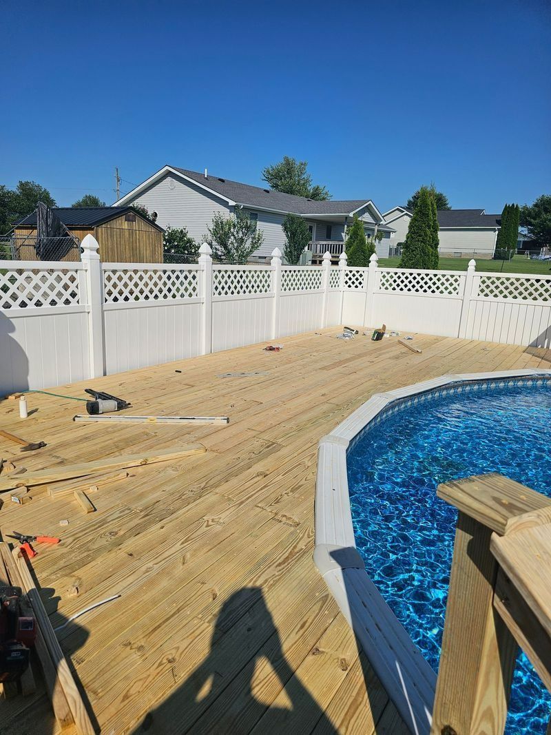 Wooden deck under construction next to an above-ground pool and a white fence, sunny day.