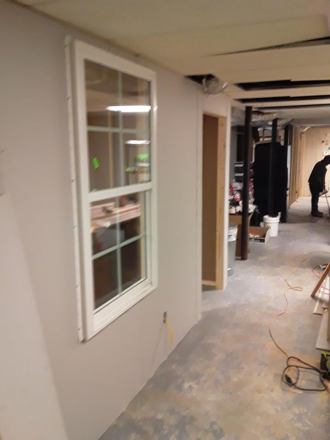 Window in room under construction; drywall, open doorway, concrete floor, person working in background.