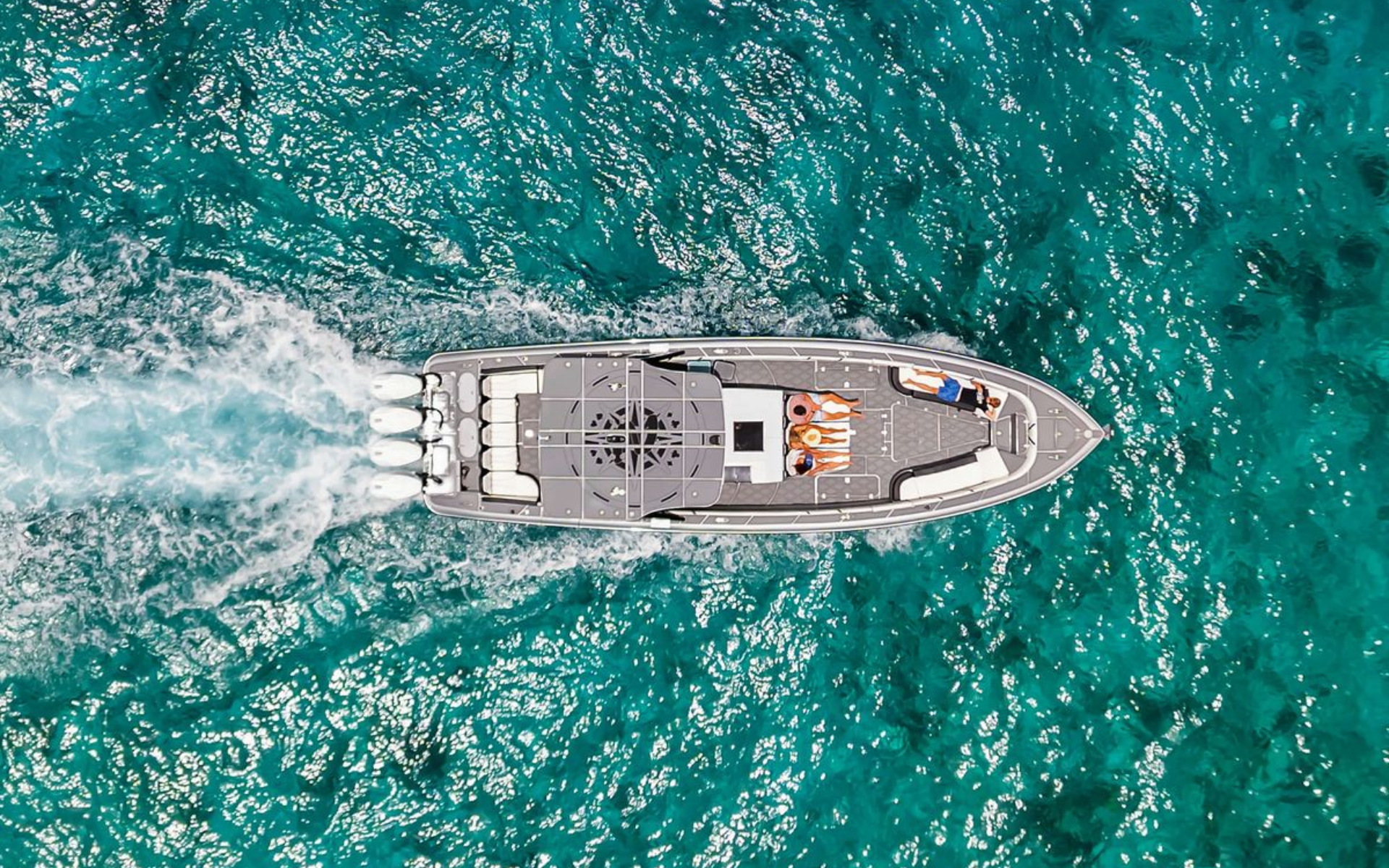 A boat speeding across turquoise water, creating a white wake. Two people sunbathe on the deck.