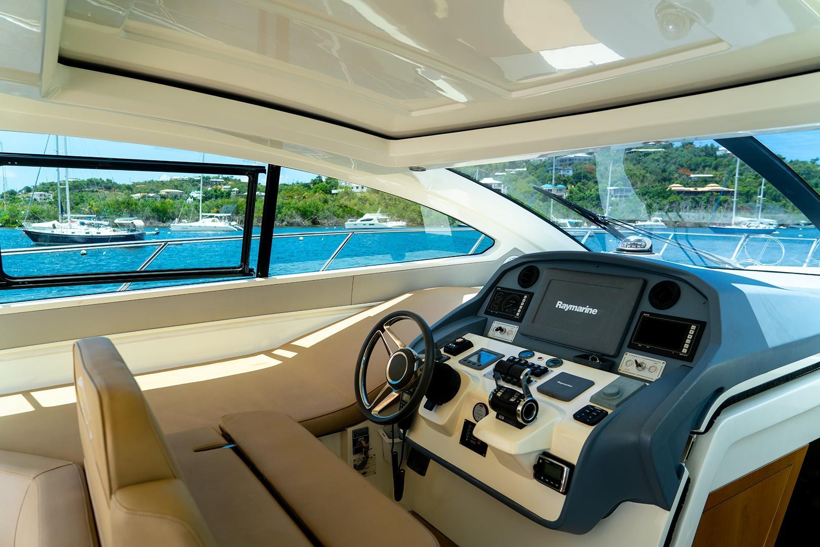 Boat cockpit interior, steering wheel, control panel, view of water and boats.