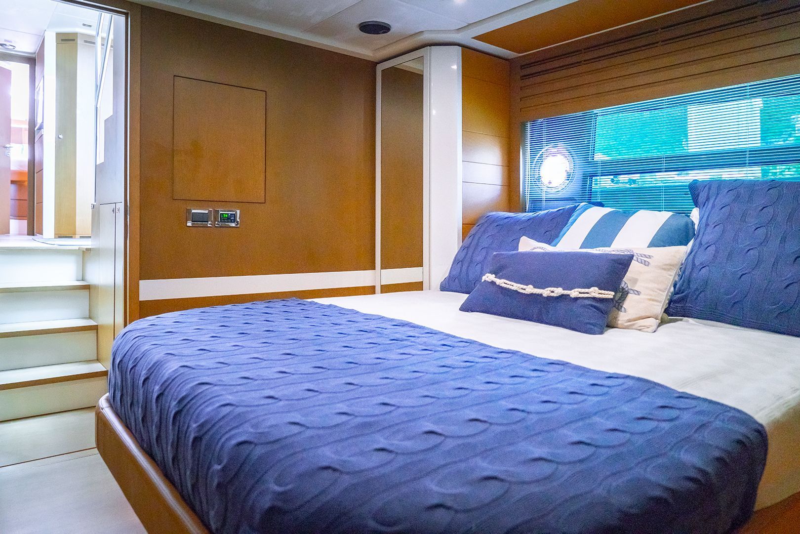 Bedroom on a yacht with a blue bedspread and pillows. Wooden walls, steps, and a window with blue and white stripes.
