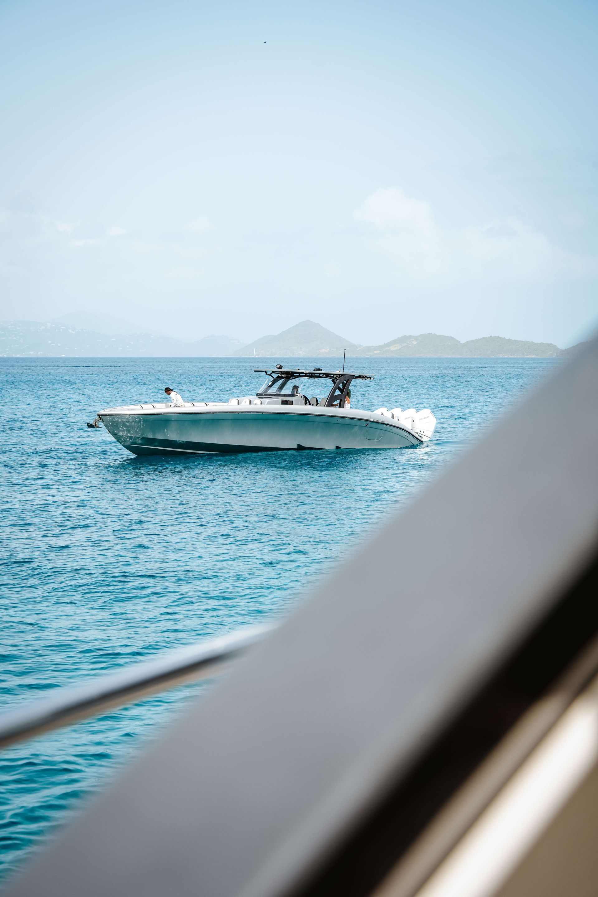 White motorboat on blue ocean, near a dark gray railing. Mountains visible in the distance.