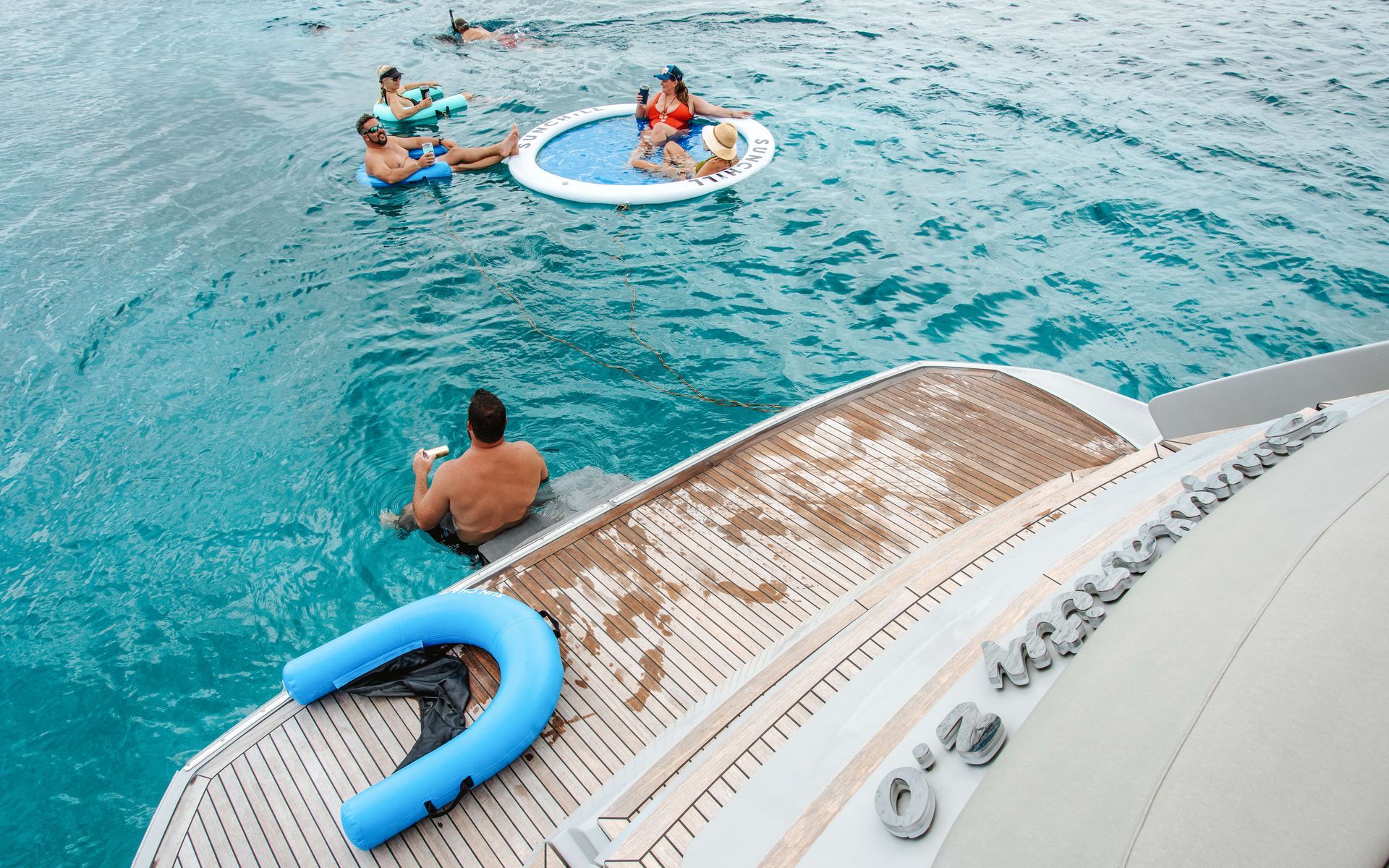 People swimming near a yacht, some in an inflatable pool and on tubes, turquoise water.