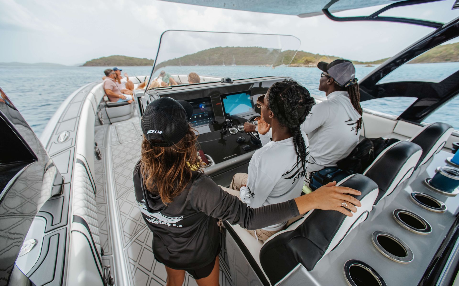 People on a speedboat, operating controls; others seated. Ocean and island backdrop, sunny.