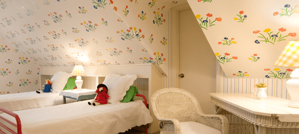 Cream wallpaper patterned with colourful flowers in a child's bedroom with rag dolls on white twin beds